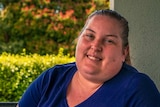 A woman in a blue t-shirt smiles for a photo on her porch, a green hedge behind her.