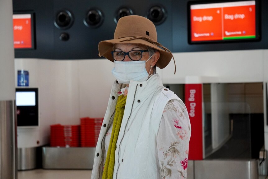 A woman wearing a brown hat, glasses and a face mask stands with a bag drop in the background.