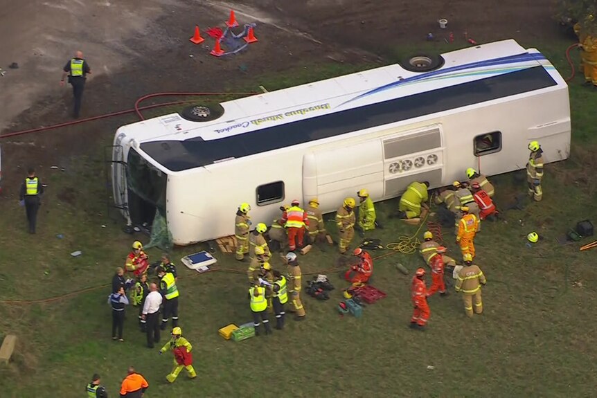A white bus on its side surrounded by emergency services personnel in high vis.