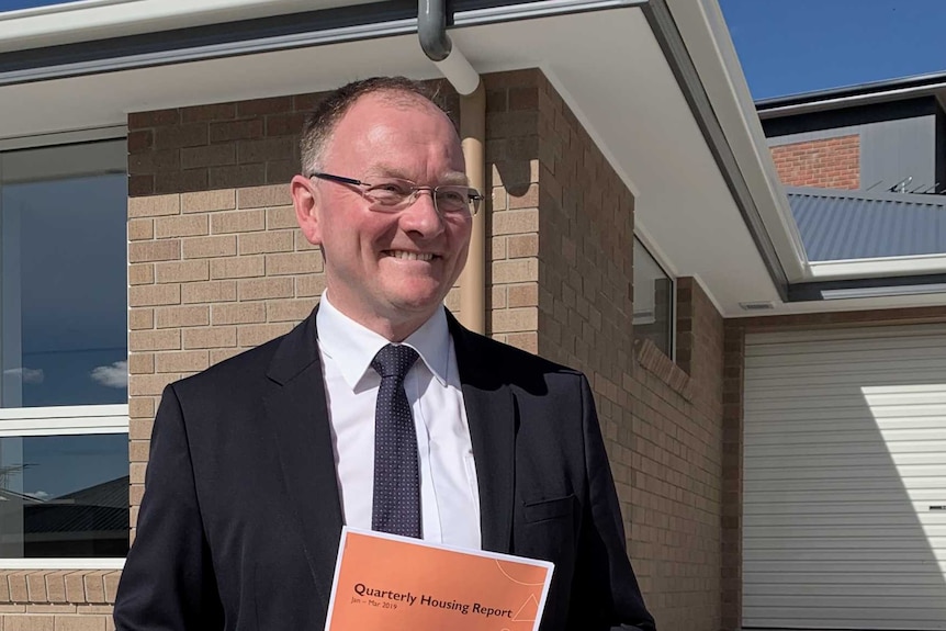 Tasmanian Housing Minister Roger Jaensch holding a booklet in front of a house.