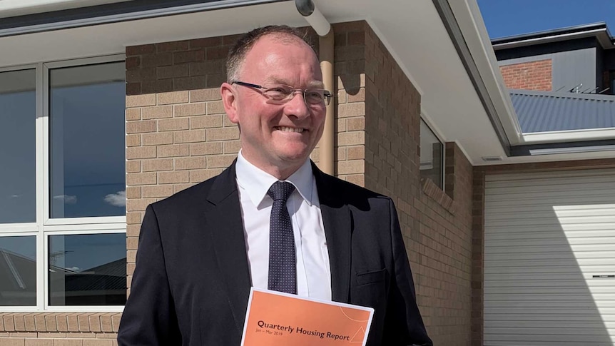 Tasmanian Housing Minister Roger Jaensch holding a booklet in front of a house.