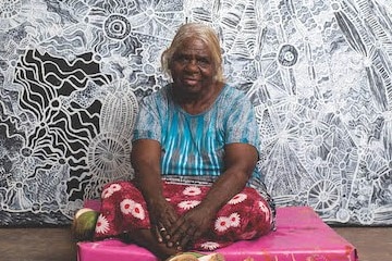 An older Aboriginal woman with grey hair, the artist Betty Muffler, sitting in front of a black and white painting