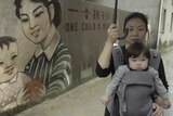 Colour still of Nanfu Wang with her child in a carrier, holding umbrella in front of mural in 2019 documentary One Child Nation.