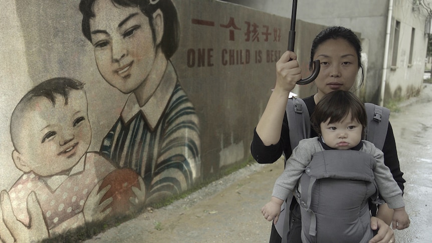 Colour still of Nanfu Wang with her child in a carrier, holding umbrella in front of mural in 2019 documentary One Child Nation.