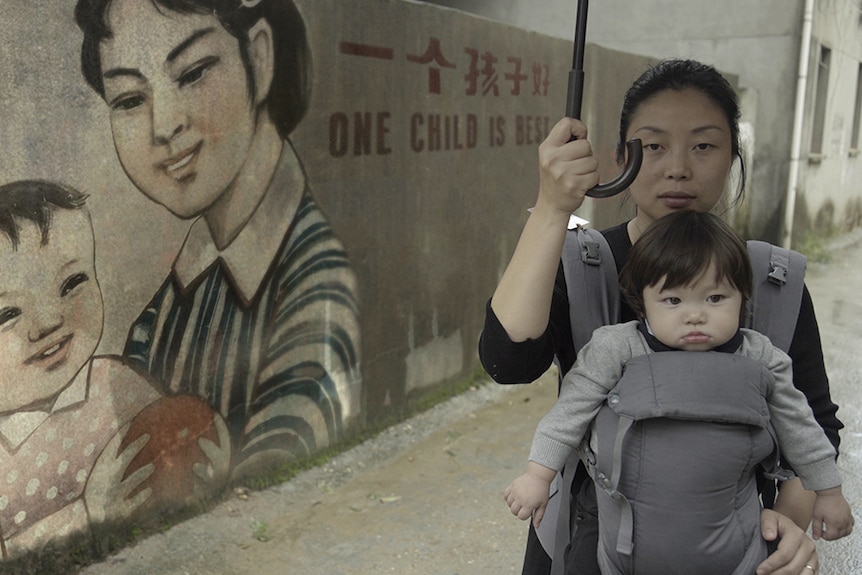 Colour still of Nanfu Wang with her child in a carrier, holding umbrella in front of mural in 2019 documentary One Child Nation.