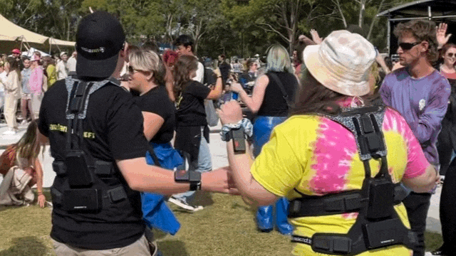Two people wearing futuristic vests dance at a festival with their backs away from the camera. 