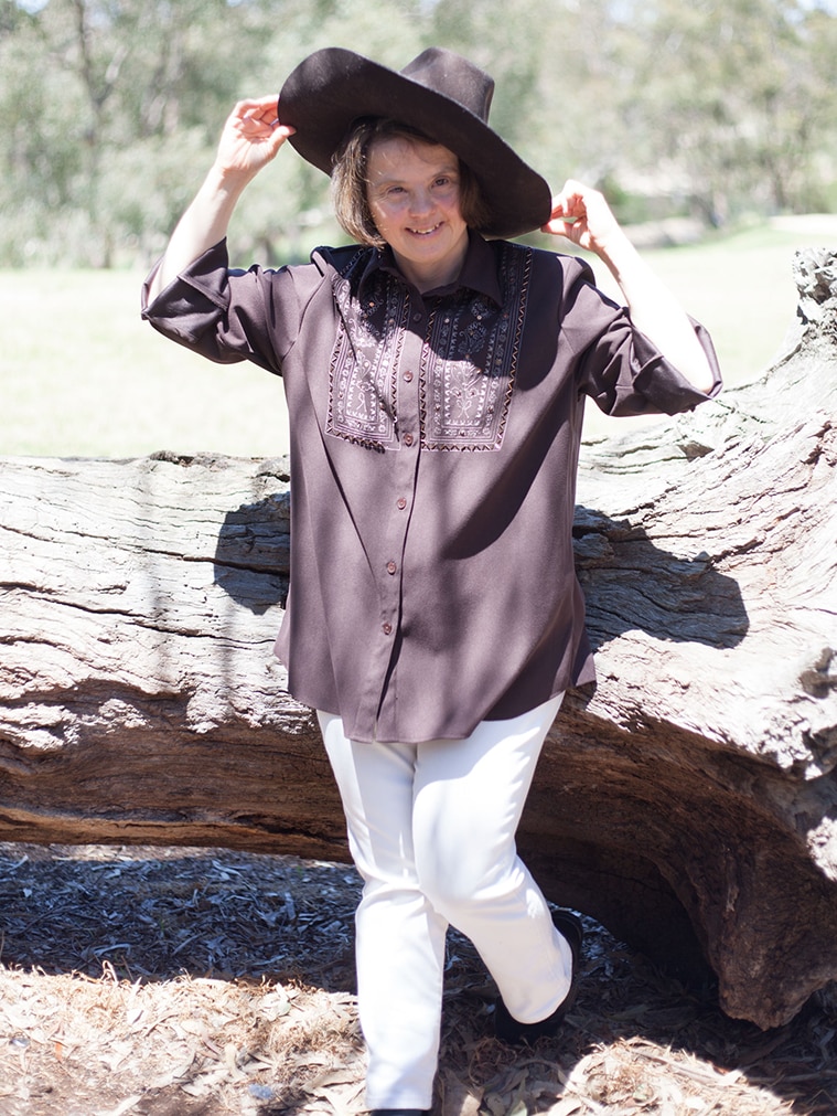 A young woman stands in front of a tree stump in the country, wearing an Akubra.