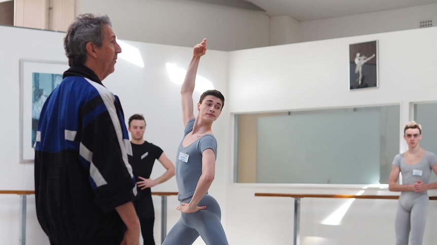 A ballet dancer in grey tights is watched by his teacher.