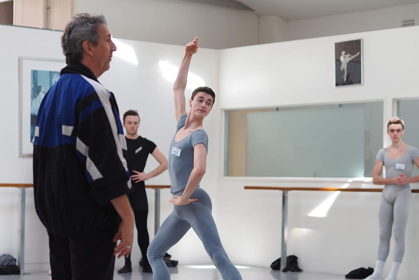 A ballet dancer in grey tights is watched by his teacher.