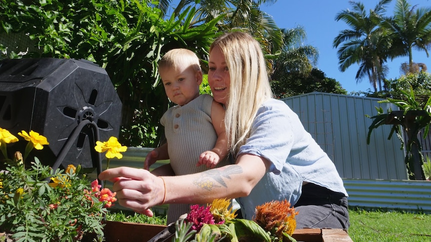 A mother and her toddler child in the garden on a sunny day, the mum is smiling and leaning past her child to pick a flower 