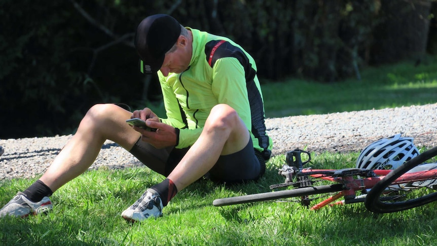 A cyclist sitting on grass looking at his phone.