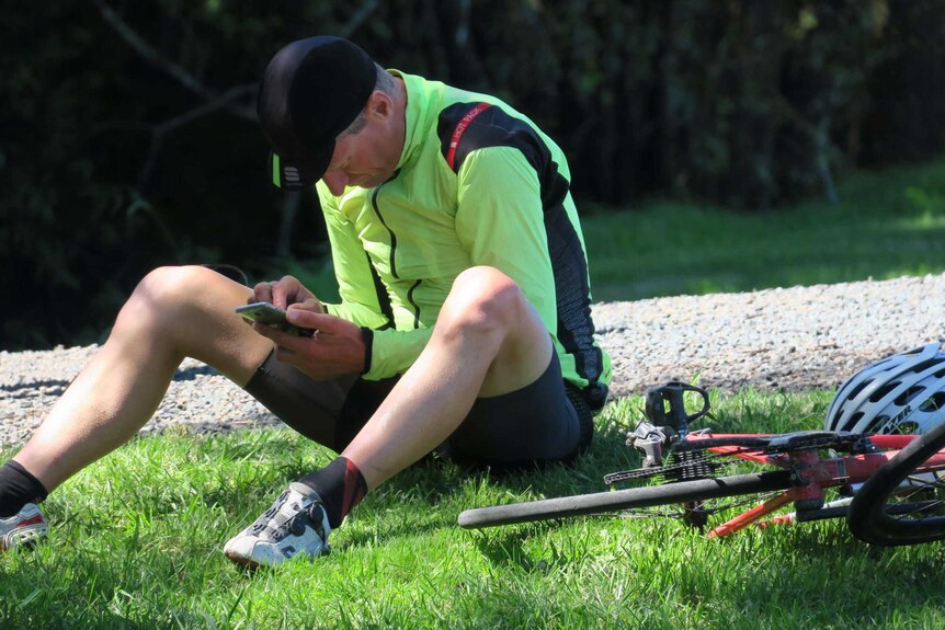 A cyclist sitting on grass looking at his phone.
