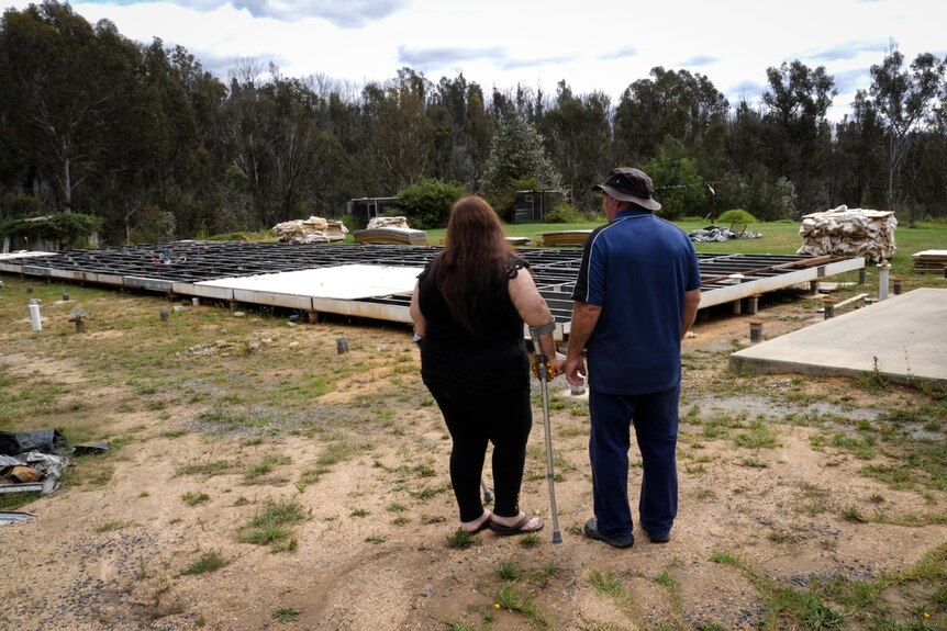 Wide shot from behind of man stand with woman using walking sticks looking at house foundations