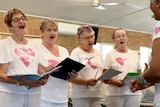 A group of women in pink headbands and white shirts sing.