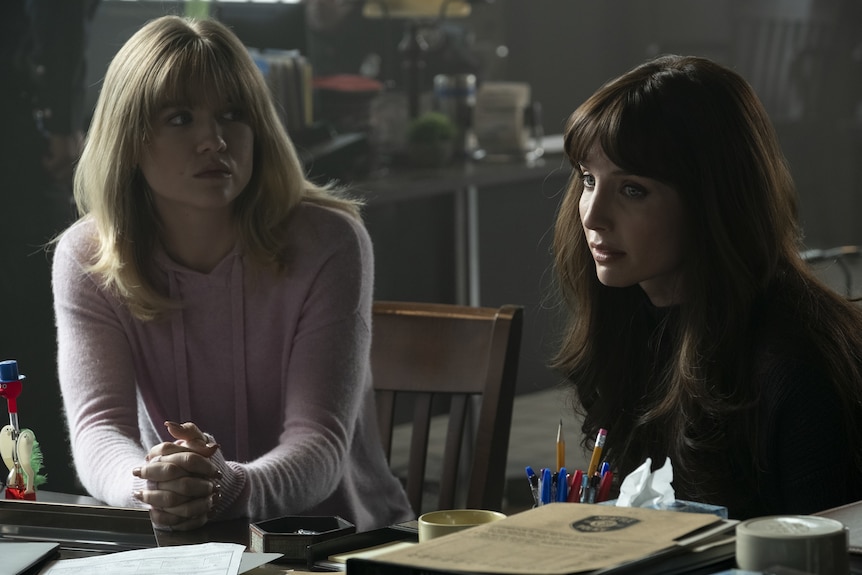 Two young women sit at a desk looking concerned, one with blonde hair and the other brown