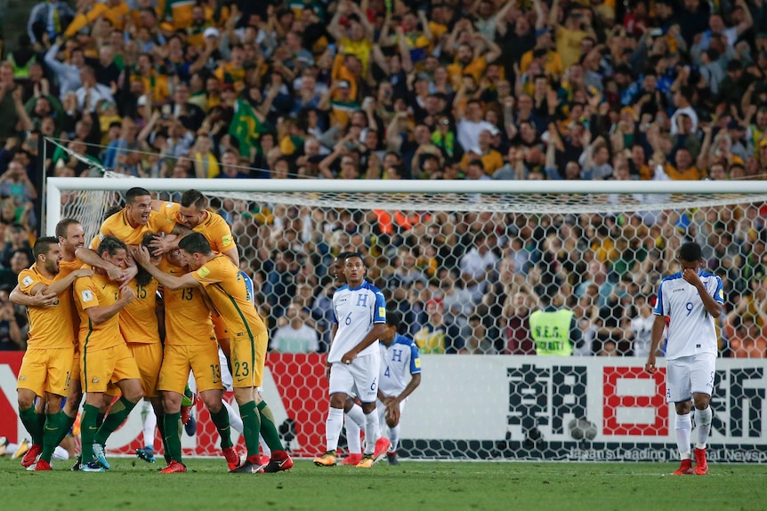 A group of Socceroos players pile on top of a teammate in celebration of a goal, while Honduras players look dejected.