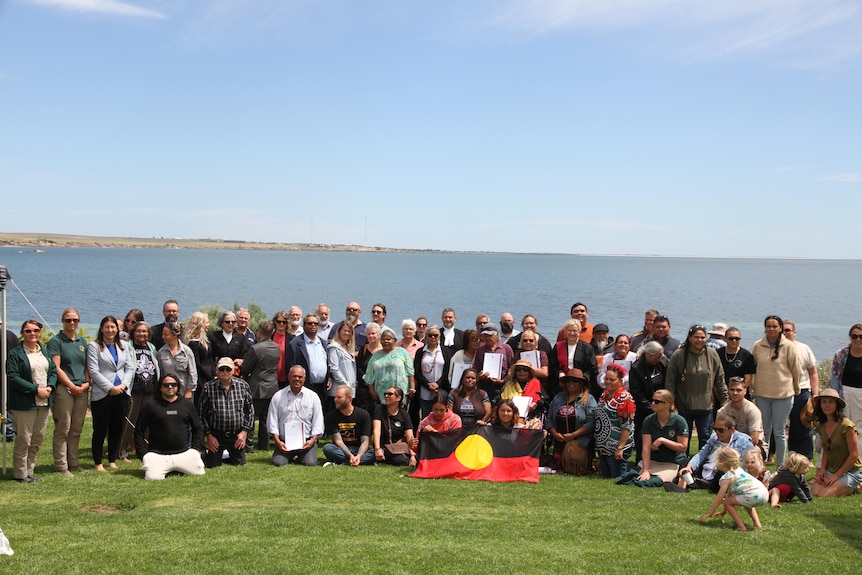 a group of about 50 people smile at a camera on a foreshore with the Aboriginal flag centred.