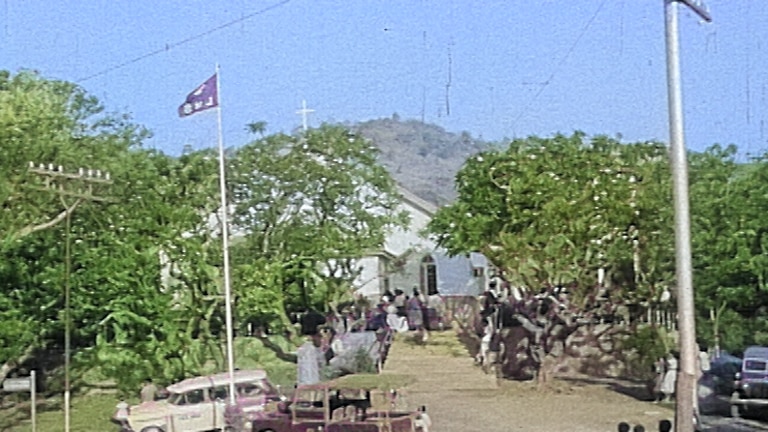 A wide street scene with a church building in the background, then a flag pole, some cars and people about the place.
