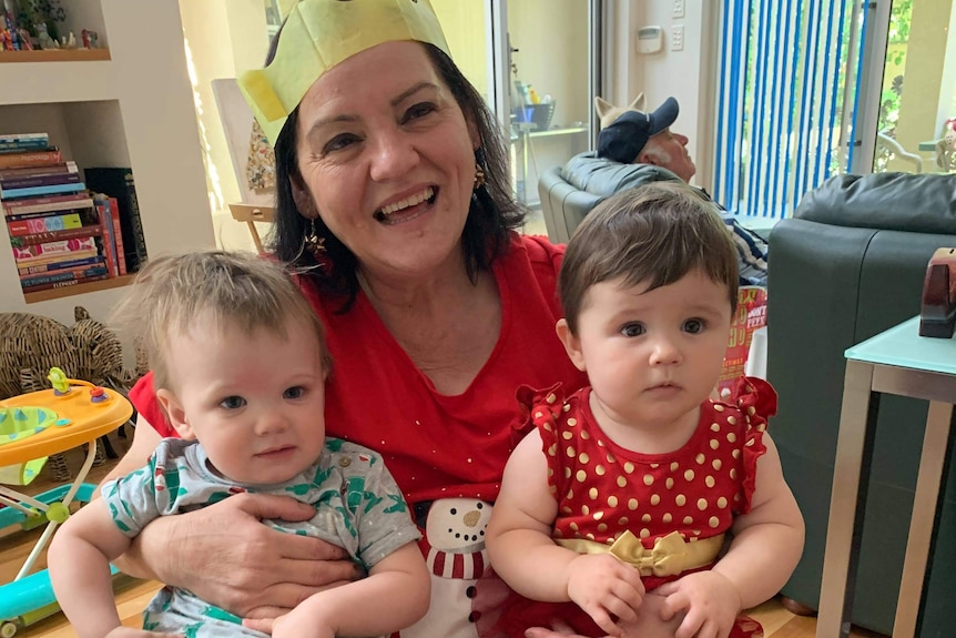 A woman in a yellow paper Christmas hat holds two babies on her lap 