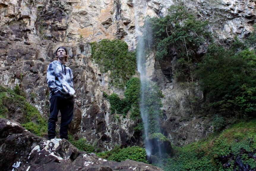 A young man wearing a hoodie and beanie stands by a waterfall during a hike
