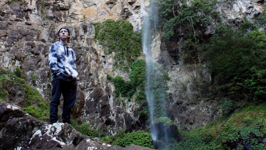 A young man wearing a hoodie and beanie stands by a waterfall during a hike