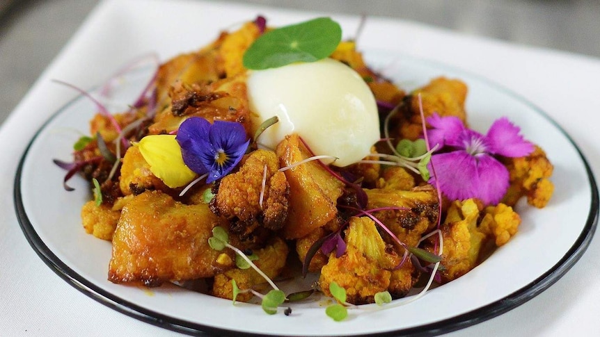 Aloo gobi dish in white plate, including fried-yellow cauliflower and potato with edible flowers on top.