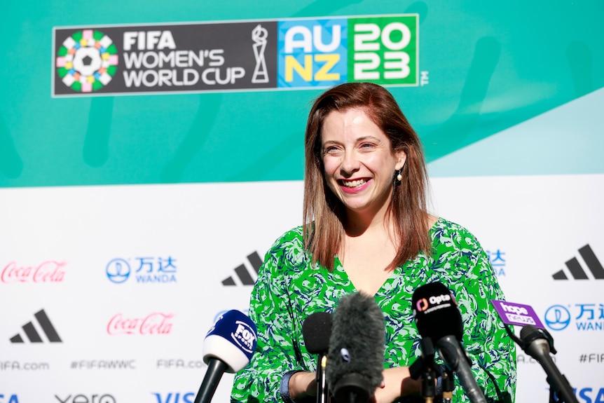 A woman in a green dress smiles in front of some microphones during an event for a sports tournament.