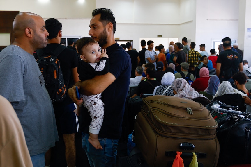 A man carrying a baby with suitcases behind talking to another man amongst a crowd of people