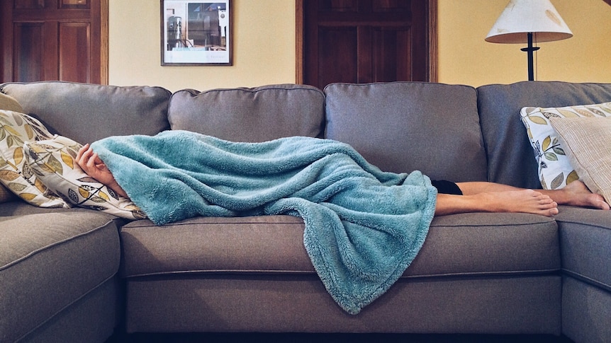 A person lays across a grey couch with a blue blanket over their face and body. 