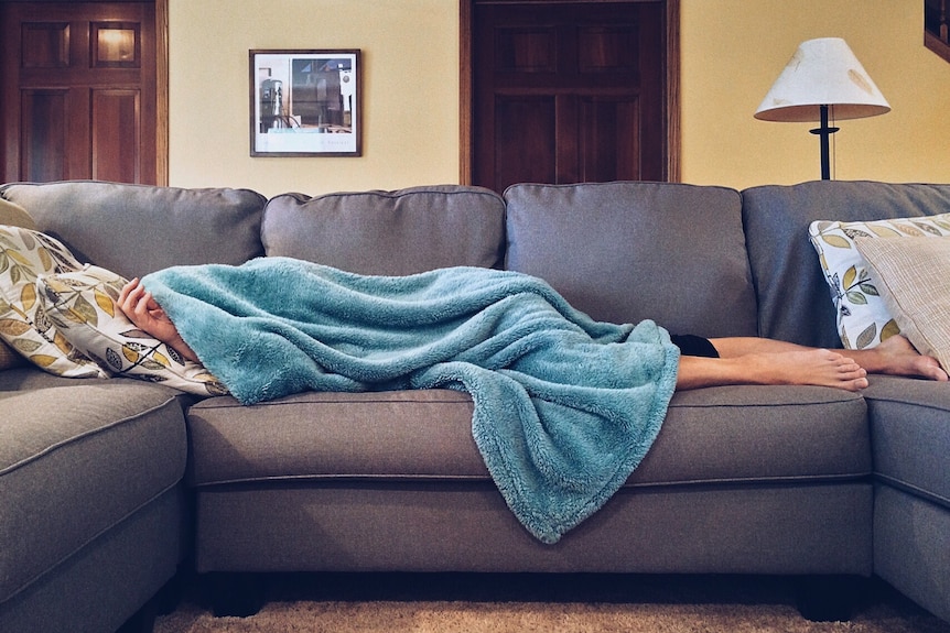 A person lays across a grey couch with a blue blanket over their face and body. 