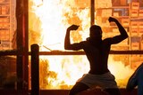 The silhouette of a man flexing both bicep muscles can be seen in front of a roaring fire inside a building.