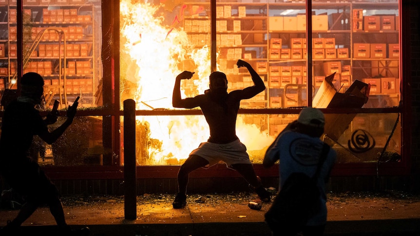 The silhouette of a man flexing both bicep muscles can be seen in front of a roaring fire inside a building.