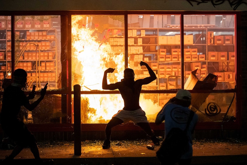 The silhouette of a man flexing both bicep muscles can be seen in front of a roaring fire inside a building.