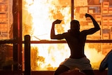 The silhouette of a man flexing both bicep muscles can be seen in front of a roaring fire inside a building.