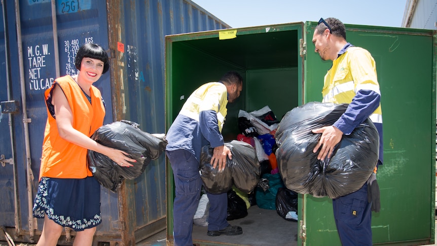 A woman and two men load bags full of linen and towels into a shipping container.