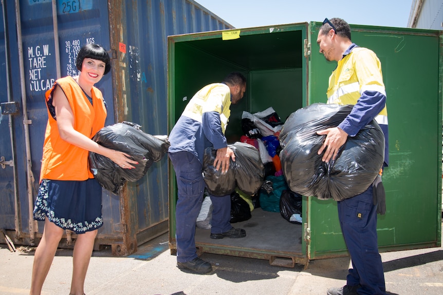 A woman and two men load bags full of linen and towels into a shipping container.