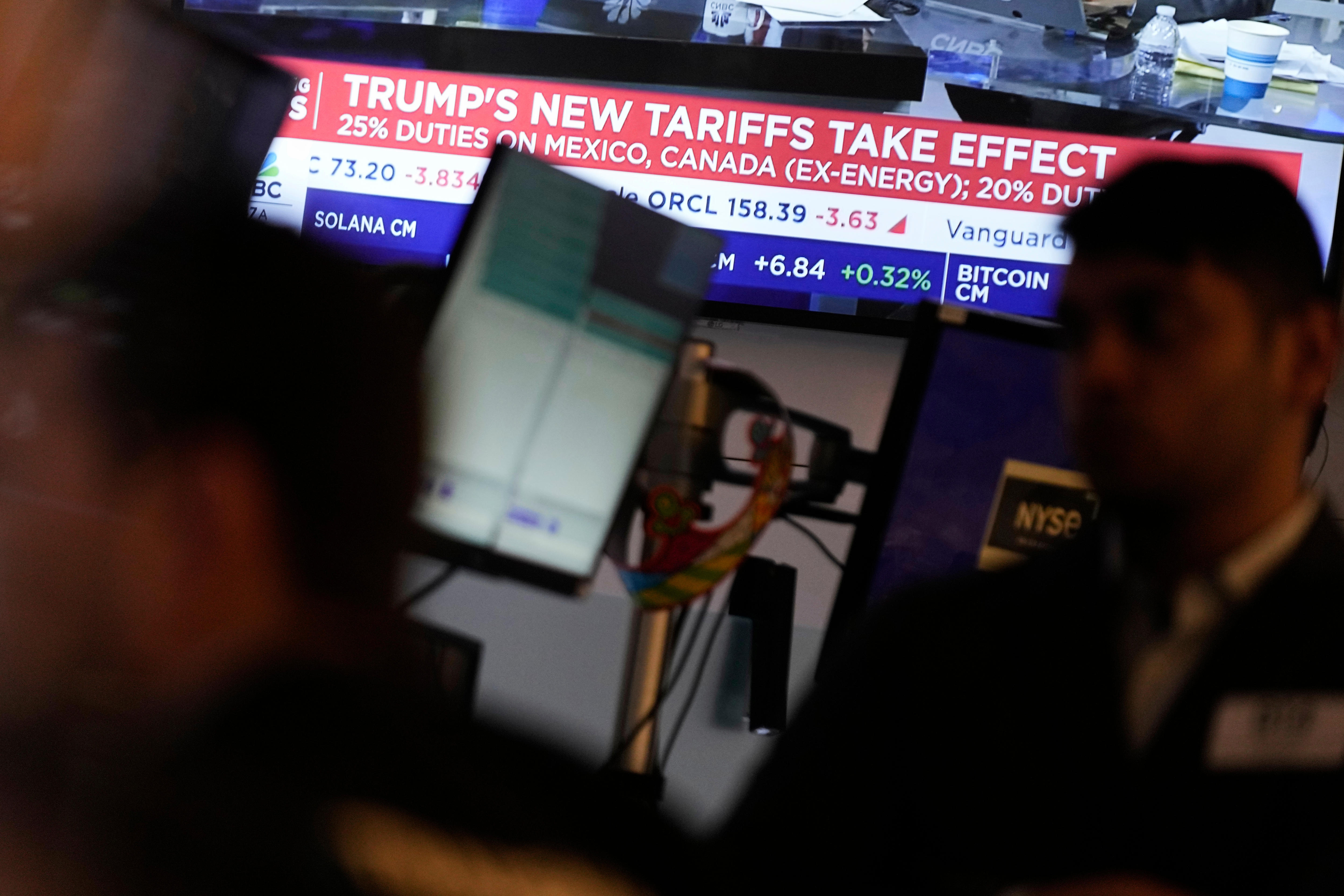 A stock market trading floor, with a TV behind them showing a headline about Trump's tariffs taking effect.