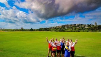 A group stands in the middle of a green football oval raising their thumbs up.