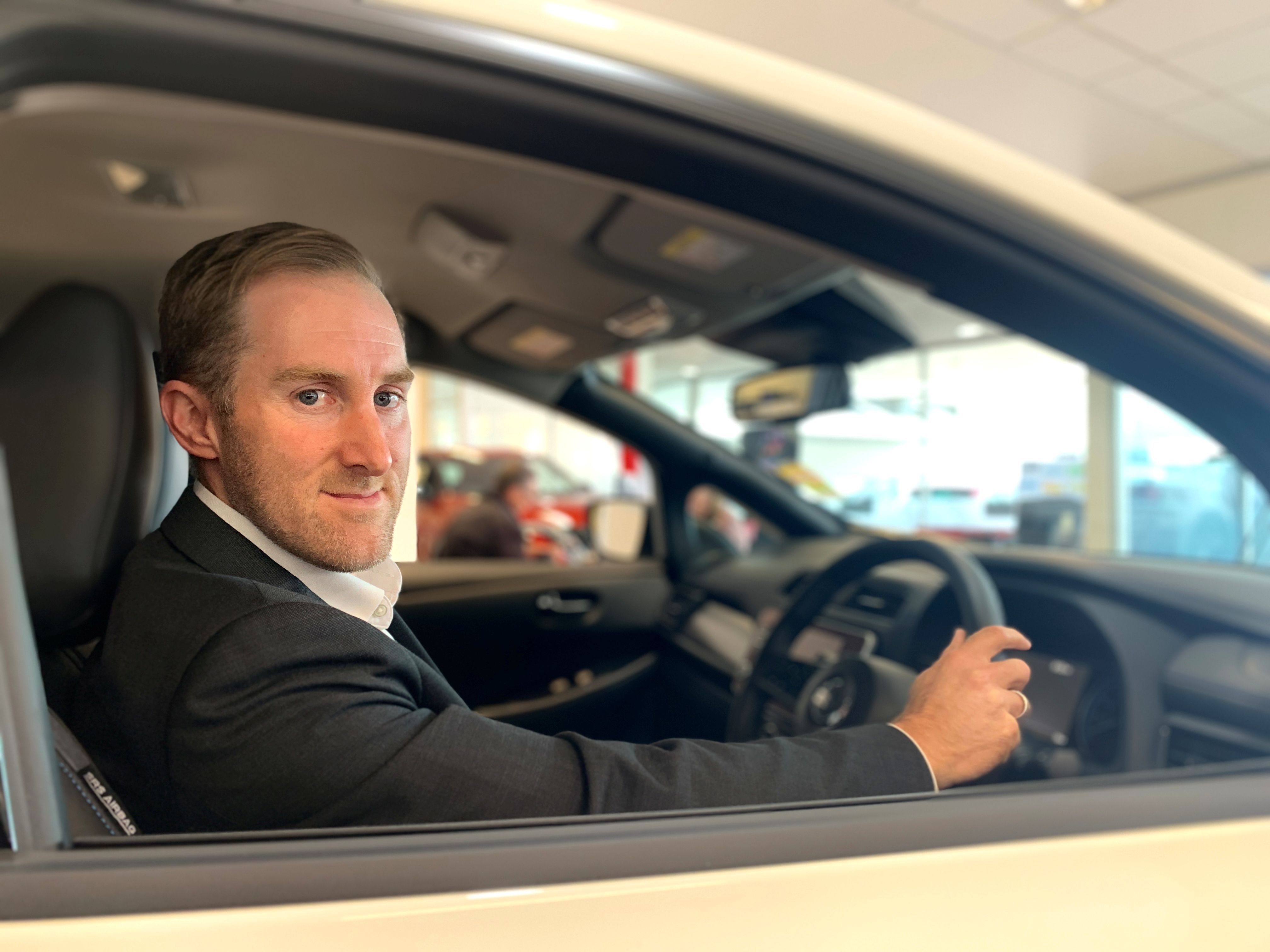 A Nissan employee sits in a parked car holding the steering wheel and looking at the camera