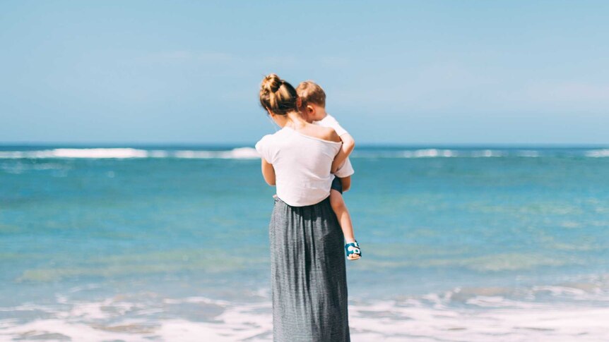 a woman wearing a long skirt stands on the beach, holding a small child