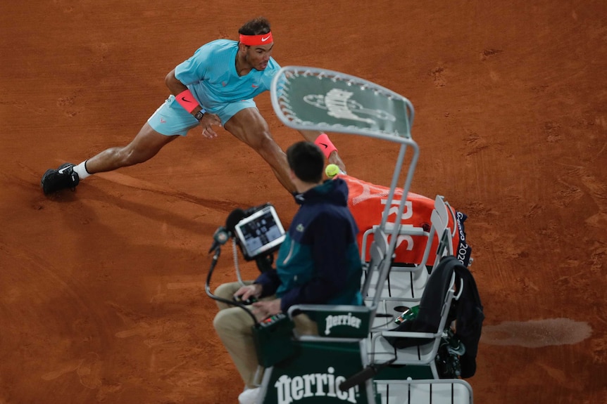 A tennis player slides across the clay to return tennis ball as the umpire watches from his chair.