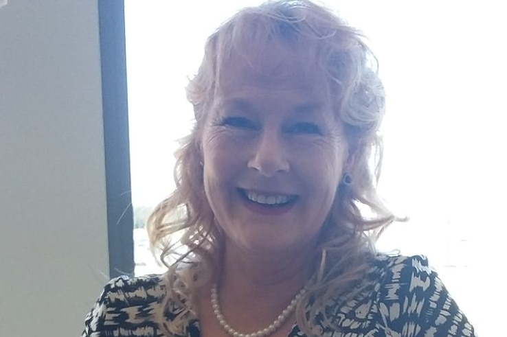 A middle-aged woman with blonde curly hair and a pearl necklace smiles broadly.