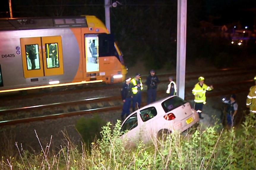A train line with a train and a car on a slope nearby with emergency services standing in a group.