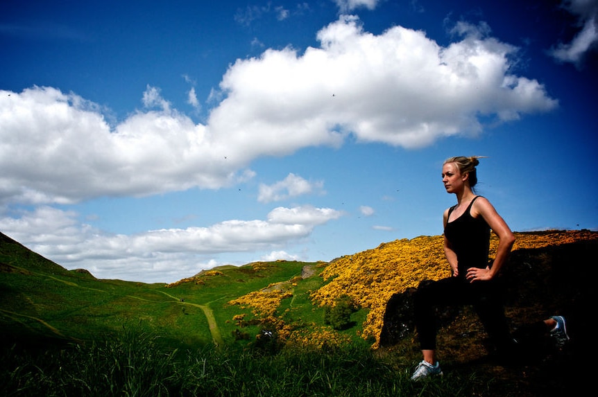 A woman does lunges in a lush green field in the middle of mountains in the edinburgh countryside