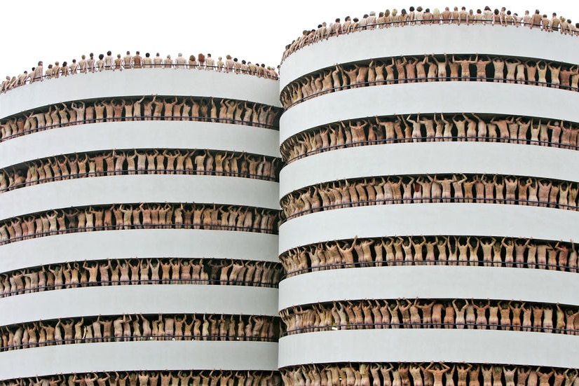 Naked models pose on multiple balconies around two white cylindrical buildings.