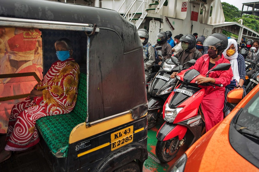 A woman with a face mask, wearing a colourful saree, sits in an auto-rickshaw with others on scooters behind her.