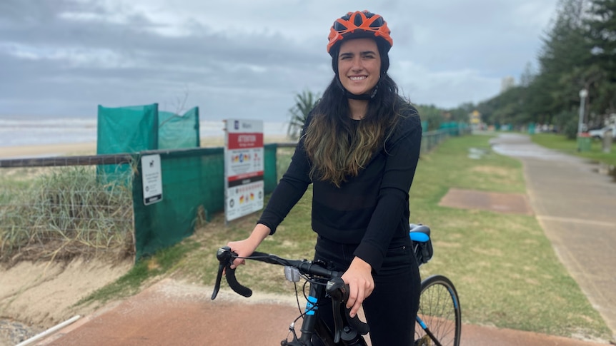 A woman with long brunette hair is seated on a bike beside the ocean, smiling.