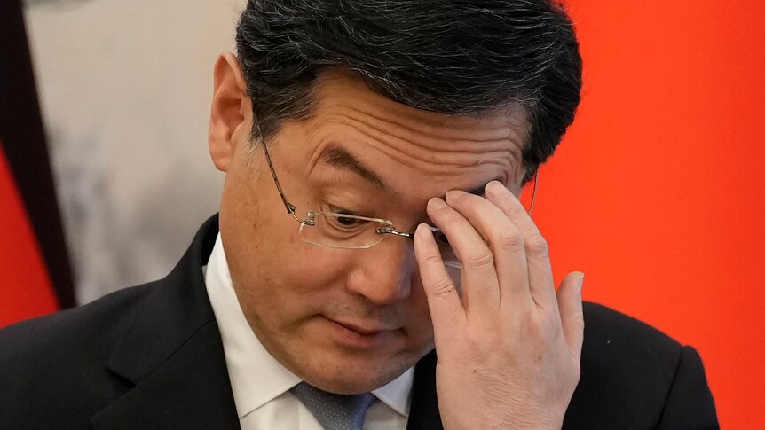 Former Chinese foreign minister Qin Gang scratches his left eyebrow with his left hand as he looks down through spectacles.