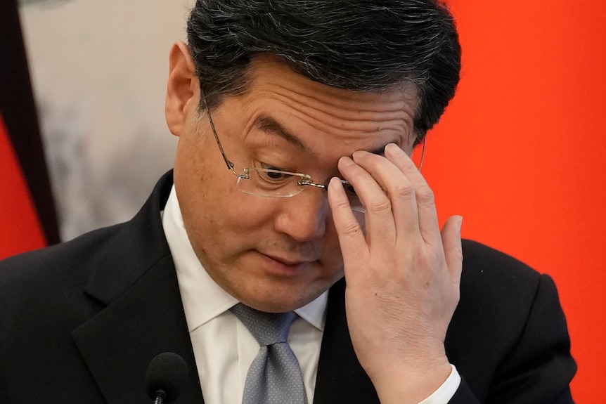 Former Chinese foreign minister Qin Gang scratches his left eyebrow with his left hand as he looks down through spectacles.