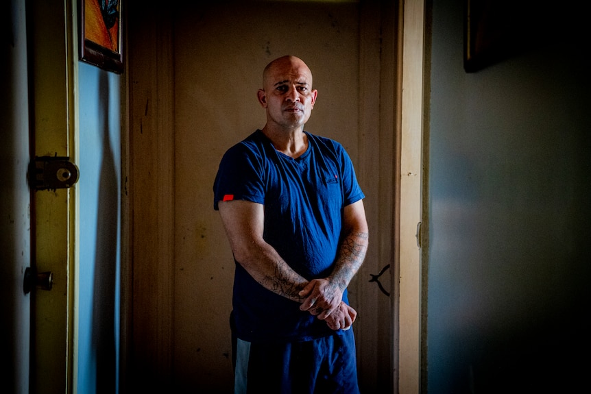 Man standing in a hallway wearing a blue shirt and his arms in front of his waist.